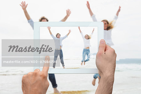 Hand holding tablet pc showing cheerful young couple jumping at beach