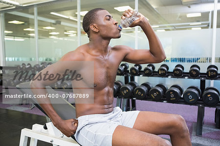 Side view of a young shirtless man drinking water at the gym