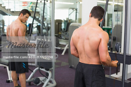 Rear view of a shirtless young muscular man using triceps pull down in gym