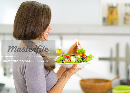Young housewife eating greek salad in kitchen. rear view