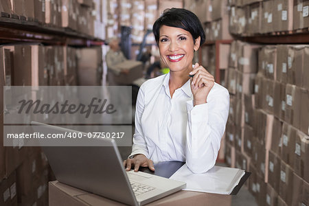 Pretty warehouse manager smiling at camera using laptop in a large warehouse