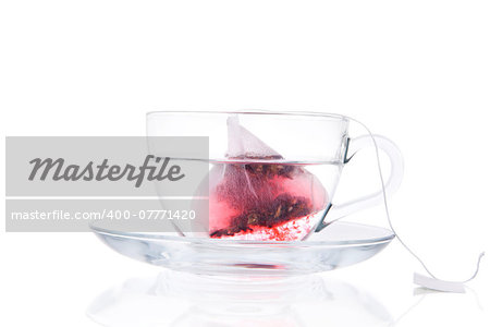 Tea Bag In Transparent Cup With Water Isolated On White Background. Red fruit tea slowly dissolving in hot water. Healthy drinking.