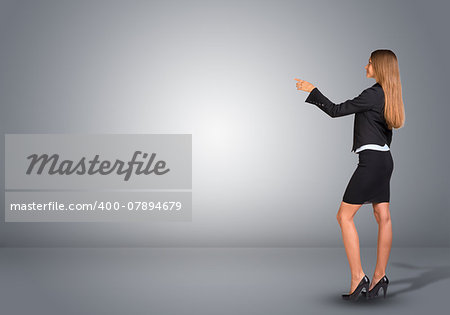 Businesswoman in suit standing in an empty gray room and pointing finger at wall. Rear view