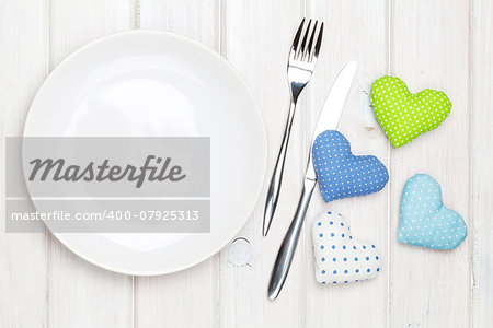 Empty plate, silverware and valentines day toy hearts. View from above over white wooden table