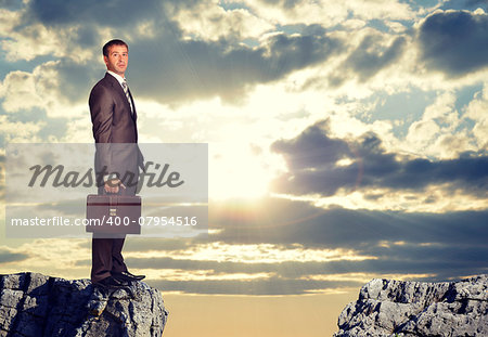 Businesswoman standing on the edge of rock gap, looking at camera as if uncertain. Sky with sun shining through clouds as backdrop