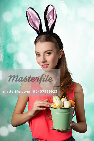 Pretty, happy blonde woman with long hair and rabbit ears on the head. She is holding bucket of little Easter eggs.