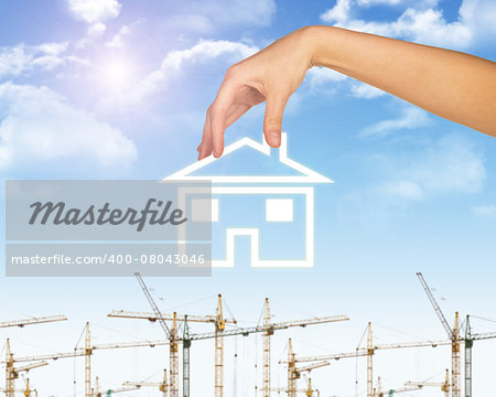 Hand holding house icon. Background of blue sky, clouds and sun, tower crane blur