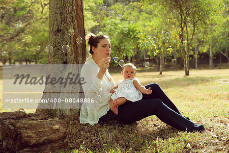 Beautiful woman sitting down tree in park and leaning on trunk with her little baby on legs, playing with her daughter blowing soap bubbles and smiling. Wide shot at sunset