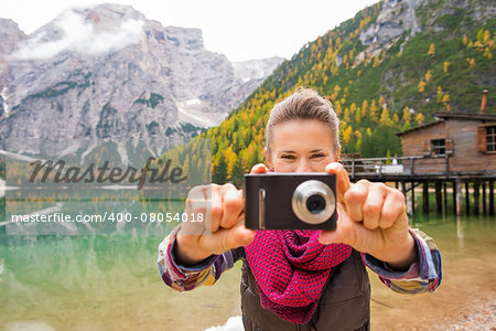 A brunette with laughing eyes is holding her digital camera up to take a photo of the viewer. We see only her smiling eyes and half her face. She is wearing outdoor gear. The warm colours in the background, including the rustic wooden pier and small house, and the Dolomites, are reflected in the still lake water.