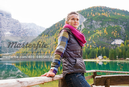 A pensive brunette hiker at Lake Bries wearing outdoor gear is leaning with her hands up against a wooden railing, smiling gently. The still water in the background provides a mirror image of the mountains and trees.