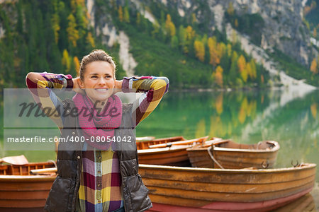 Wearing outdoor gear, a brunette woman hiker relaxes, hands behind her head. She is happy and content. In the background, wooden boats float on the water. Autumn colours and trees are reflecting off the still water.