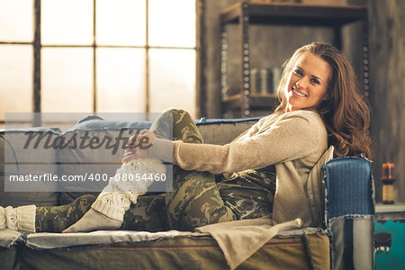 An elegant brunette smiling woman in comfortable casual clothing, leggings, and a cardigan is relaxing on a loft sofa. Industrial chic ambiance and cozy atmosphere.
