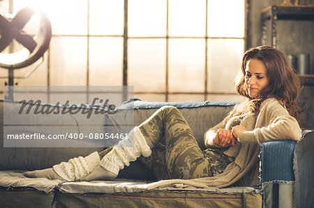 An elegant brunette woman smiling and looking down is wearing comfortable, casual clothing, leggings, and a cardigan and is relaxing on a loft sofa. Industrial chic ambiance and cozy atmosphere.