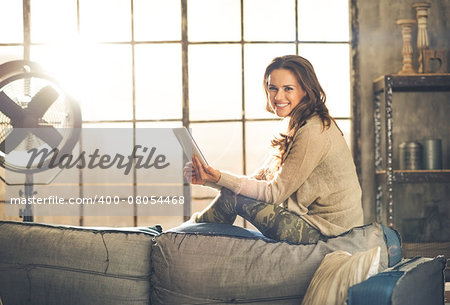 A brunette woman is smiling, looking up from her  tablet PC while sitting on the back of a sofa. Industrial chic ambiance and cozy atmosphere, sunlight is streaming through the loft window.