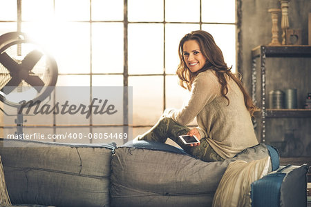 A brunette woman is smiling with phone sitting on the back of a sofa. Industrial chic ambiance and cozy atmosphere, sunlight is streaming through the loft window.