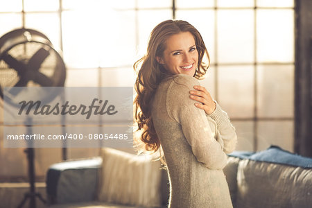 A smiling brunette woman in comfortable clothing is standing in a loft living room, hugging herself while looking over her shoulder. Urban chic loft decoration details.