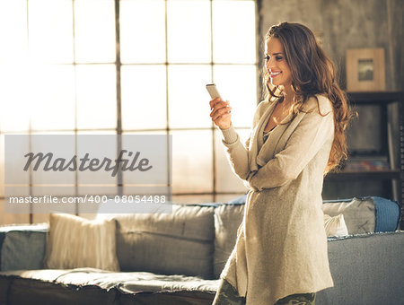 Seen in profile, a brunette woman in comfortable clothing is standing in a loft living room, looking down at her phone and smiling. Urban chic loft decoration details and window.