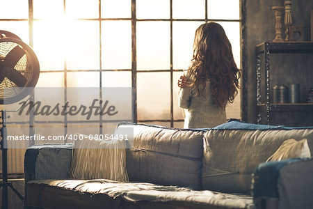 Looking away, a brunette woman in comfortable clothing is standing in a loft living room, hugging herself, looking out the window. Urban chic loft decoration details and window.