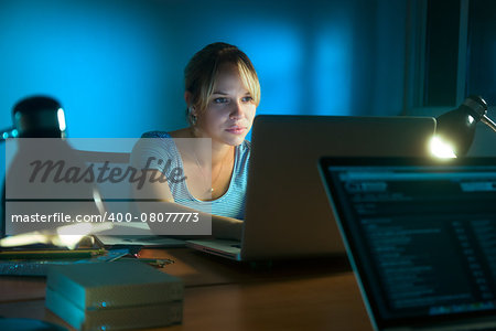 Beautiful woman working late at night in office, surfing the web and writing post on social network with laptop computer. The girl is concentrated and serious