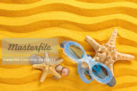 Blue swimming goggles, pebbles and seashells on yellow sand.