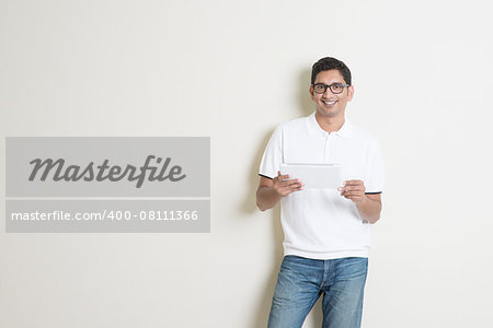Portrait of handsome Indian guy using tablet pc, standing on plain background with shadow, copy space at side.