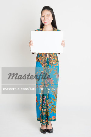 Full body portrait of Southeast Asian girl in batik dress hands holding white blank placard, standing on plain background.