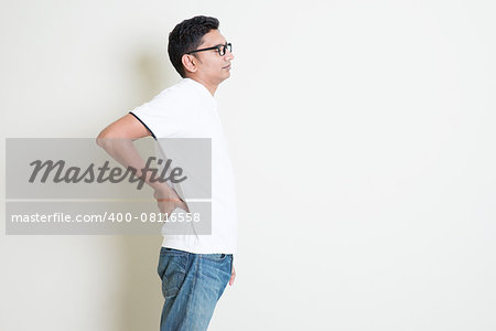 Portrait of Indian guy backache, holding spine with hand. Asian man standing on plain background with shadow and copy space. Handsome male model.