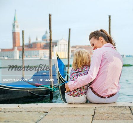 An elegant mother looks lovingly down at her daughter as she holds her close for a hug. They are happy, relaxed, and enjoying sitting by the water's edge, just spending time together.