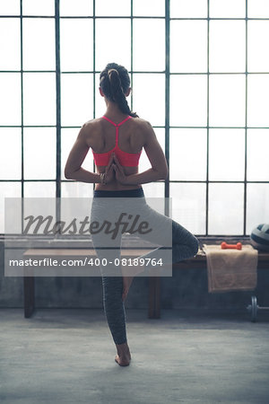 Taking things to another level, a fit, athletic woman does a yoga pose, clasping her hands behind her back and standing in tree position.