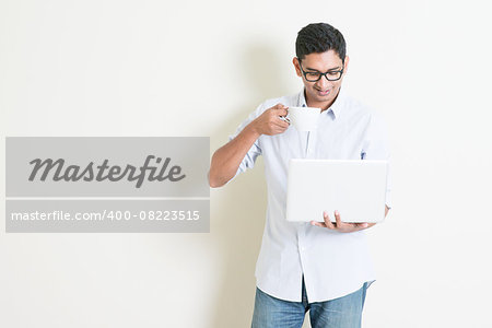 Portrait of handsome casual business Indian man drinking coffee while using laptop computer, standing on plain background with shadow, copy space at side.