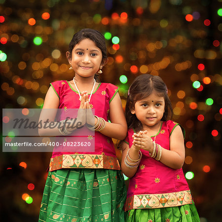 Cute Indian girls dressed in sari with folded hands representing traditional Indian greeting, standing inside a temple celebrating diwali, festival of lights.