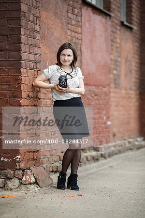 Pretty young woman with an old vintage camera in hand posing on a background of an old brick wall. Shallow depth of field. Selective focus on model. Vertical shot. Retro style photo.