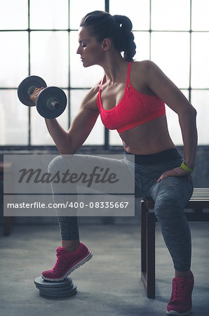 A fit, muscular woman sitting on a city loft gym bench is lifting weights, resting her elbow against her knee, curling the weights towards her as she looks down at her hand.