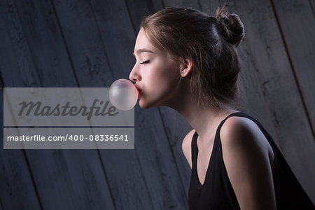 Portrait of sexual brown-haired, which inflates languidly pink bubble of chewing gum. Photographed in studio on a dark background.