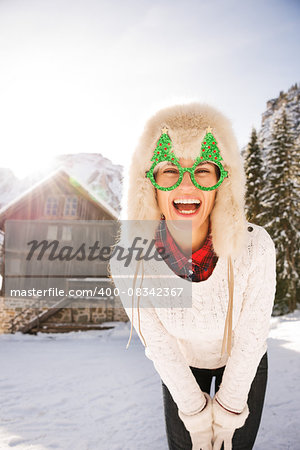 Going wild for Christmas season in a secluded spot in the countryside. Portrait of laughing young woman in funny Christmas tree glasses in the front of a cosy mountain house.