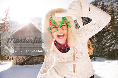 Going wild for Christmas season in a secluded spot in the countryside. Smiling young woman in funny Christmas tree glasses framing with hands in the front of a cosy mountain house.