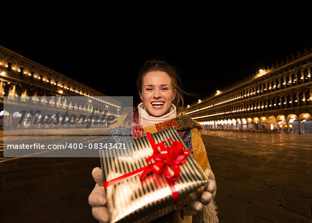 Excitement of Christmas time and allure of long-time favourite Venice, Italy. Happy woman showing Christmas gift box while standing on Piazza San Marco in the evening