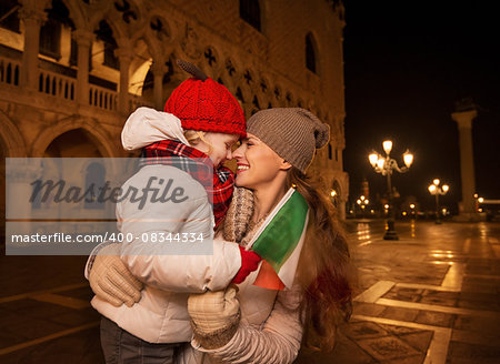 Holiday family trip to Venice, Italy can change the whole Christmas experience. Happy mother and child with Italian flag hugging while standing on Piazza San Marco in the evening. Winter Tourism