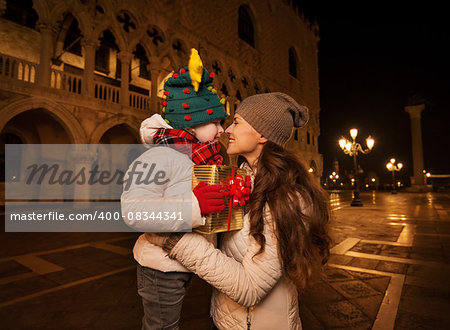 Holiday family trip to Venice, Italy can change the whole Christmas experience. Happy mother hugging child with Christmas gift box while standing on Piazza San Marco in the evening