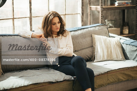 Thoughtful young woman with brown hairs is sitting on sofa in loft living room