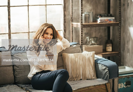 Smiling stylish brunet woman in elegant clothing sitting on divan and talking smartphone in loft apartment