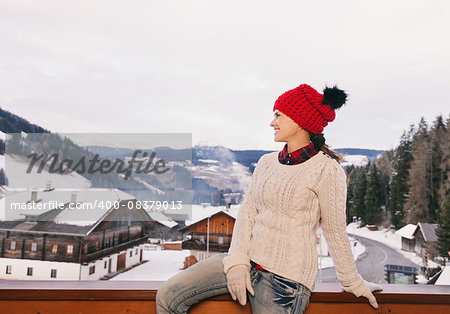 Mountain cottage hideaway brings calm and warmth into the winter season. Happy young woman on balcony overlooking the snow-capped mountains looking into the distance