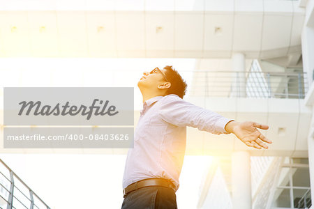 Business freedom concept. Asian businessman arms outstretched enjoying the morning breeze, morning sunlight and modern office as background.