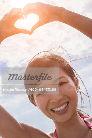 Beautiful young smiling Chinese Asian woman girl on a beach in a bikini, making a hand heart shaped finger frame