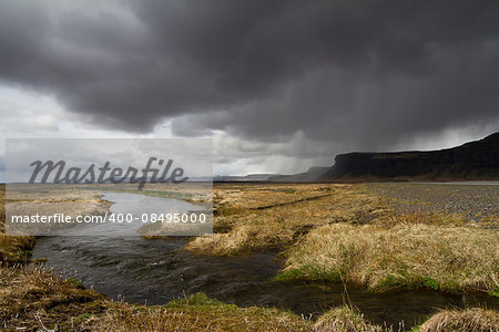 Horizontal panorama view of an Icelandic river running through the grass with rain stormy clouds above and mountains on the right side