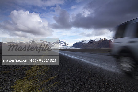 Horizontal photo of an off road car on a straight asphalt road coming from the mountains with clouds above and the Vatnajokull glacier in the background, Iceland