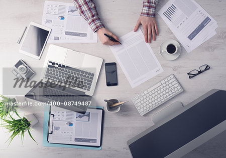 Businessman working at office desk and signing a document, computers and paperwork all around, top view