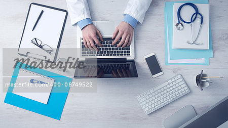 Doctor sitting at office desk and working on his laptop with medical equipment all around, top view