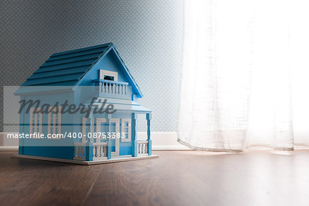 Blue wooden model house next to a window with curtain on wooden floor.