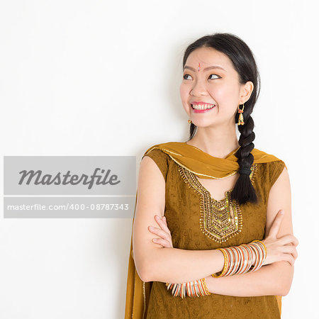 Portrait of arms crossed mixed race Indian Chinese woman in traditional Punjabi dress looking side upward, standing on plain white background.
